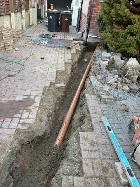 Trench along a paved path with a new orange drain pipe laid in it. One of our jobs.